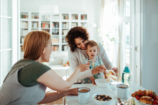 Two women feeding toddler breakfast at home kitchen