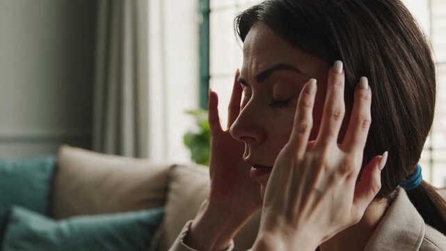 Professional businesswoman experiencing intense workplace stress, massaging temples while sitting in living room, demonstrating mental health challenges and physical exhaustion from overwork