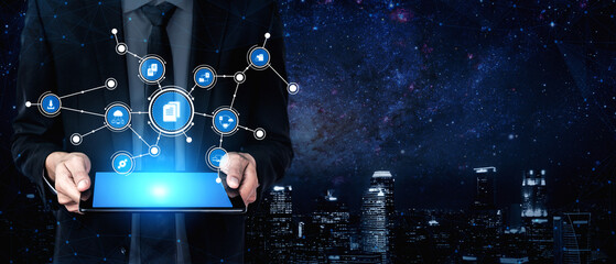 Obraz premium A professional male in a suit holds a tablet displaying digital icons representing connectivity and data networking against a vibrant urban night skyline. Tessel