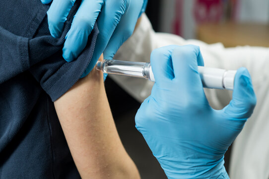 A medical professional wearing blue nitrile gloves uses an injector pen to deliver medication into the upper arm of a patient.