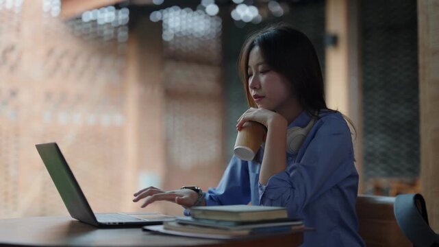Asian adult woman drinks coffee works on laptop computer reads books at modern cafe workspace freelancer student remote learning online business professional