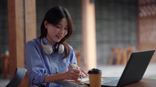 Asian adult woman studies works writing notes laptop cafe environment focused diligent learning professional education technology freelance remote