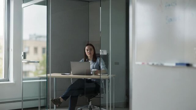 Medium shot of a compact glass pod illuminated by natural light individual concentrating on laptop beside blurred whiteboard inside for calm solo study.