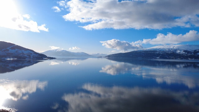 Aerial drone view of calm Norwegian fjord reflecting snowy mountains and dramatic clouds under blue sky. Scandinavian winter landscape with mirror water surface and distant mountain range.