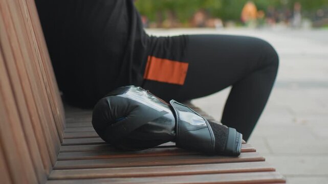 white man seated on wooden bench removing gloves and grabbing water bottle, orange shorts patch visible, stretching leg and breathing deeply, park background with distant pedestrians, relaxed recovery