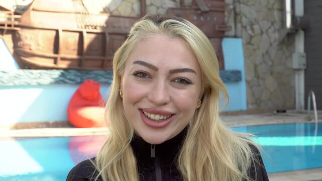 Happy young blonde woman with a radiant smile looking at camera while standing by a swimming pool at a luxury villa or resort during her summer vacation holidays outdoors at daytime