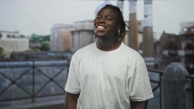Man smiling with face and teeth visible, wearing white t shirt, standing on old town street in front of stone railing; joy urban youth.