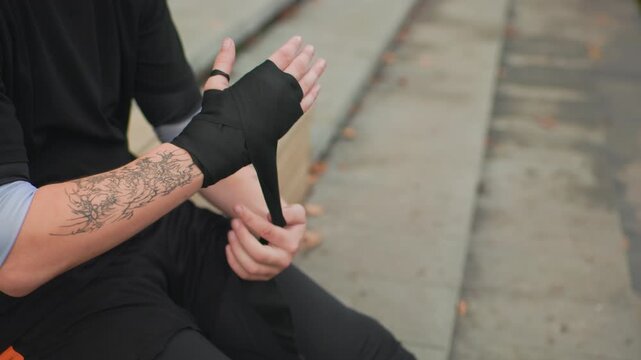 tattooed caucasian man wrapping hand bandage on steps, sitting on concrete stairs wearing black apparel, careful motions securing wrist wrap, closeup of forearm tattoo and knuckles, gritty urban
