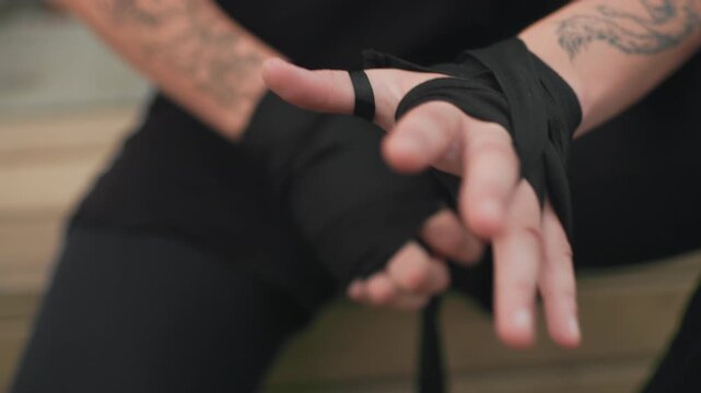 caucasian MMA fighter securing black hand wraps on bench, closeup of tattooed forearms and precise fingers threading strap, calm tactical routine before sparring or match, focused technique