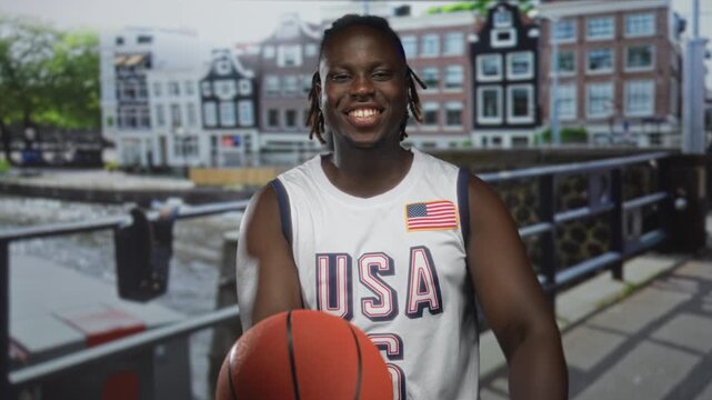 Man points finger to basketball on street beside canal guardrail, smiling with wristband and usa jersey; youth sports joy.