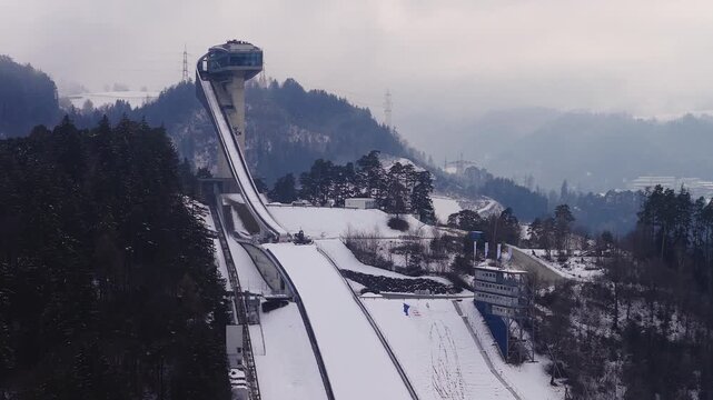 Aerial view shows Bergisel Ski Jump in Innsbruck, Austria, with curved tower, long inrun, empty landing hill, snowcat, judges platform with flags, and misty ridgelines.