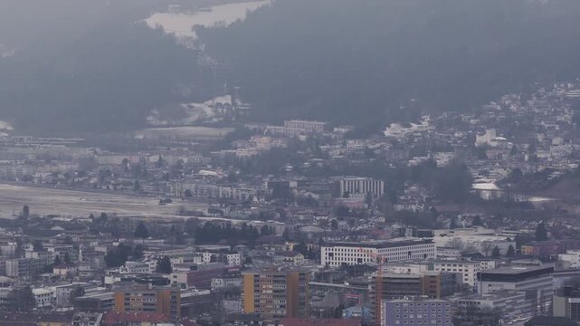 Aerial view of Innsbruck, Austria shows Nordkette slopes and Kranebitten airport as an airliner makes final approach, with winter light, haze, housing and cranes visible.