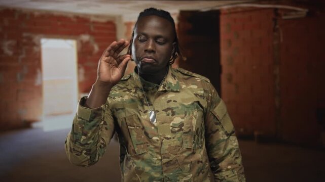 Man soldier in camouflage uniform touching his face and pinching nose with hand, dogtag visible, standing in unfinished building; duty determination.