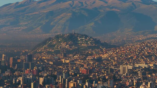 Telephoto drone view of El Panecillo in Quito, Ecuador from the north, showing the iconic hill and monument above the dense city under warm sunrise light.