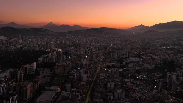 Aerial drone view of Quito, Ecuador at sunset with dense urban skyline, glowing streets, mountain silhouettes, and Cotopaxi volcano visible in the distance beneath an orange evening sky.