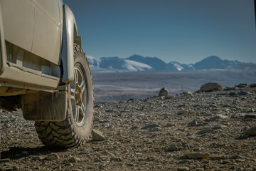 Close up of off-road 4x4 SUV wheel and mud tire on rocky edge of mountain plateau overlooking scenic snow peaks and highland valley during overland adventure journey in wilderness.  © ANDREY
