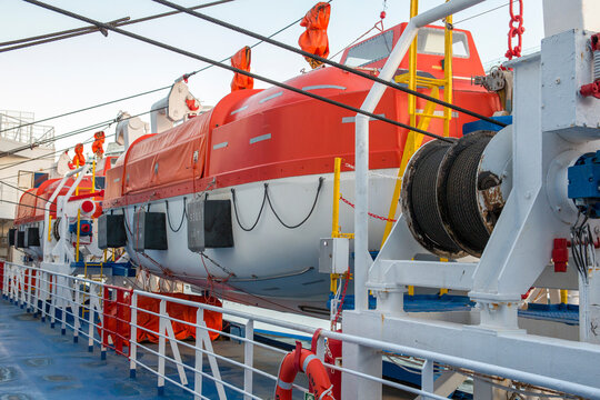 A row of orange and white lifeboats mounted on davits along the deck of a large ship. The scene shows essential maritime safety equipment, steel cables, and railings under a clear sky.