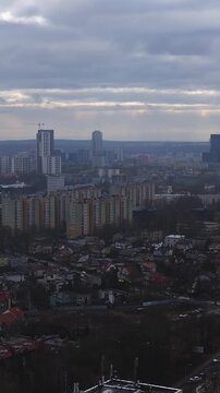 Aerial pan over Katowice, Poland shows compact houses, beige blocks, dark glass office, high rise towers, and a crane topped tower under overcast sky in vertical