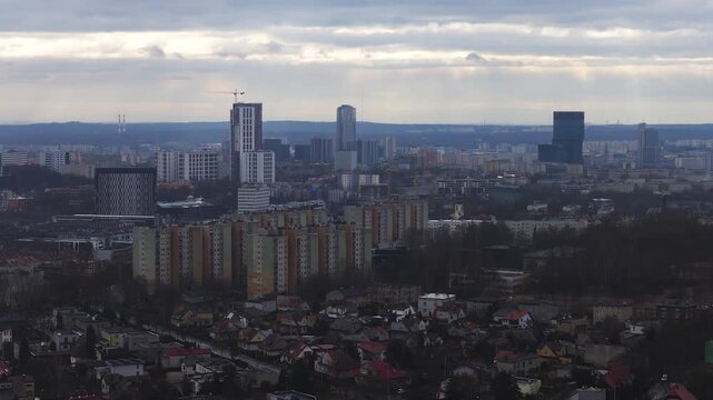 Aerial view of Katowice, Poland shows small houses, prefab blocks, new high rises, a dark glass cuboid tower, and a crane, in cool overcast light with bands of sun.