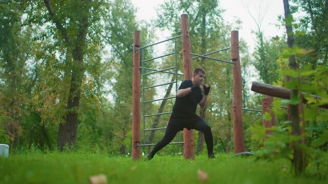 outdoor forest boxing training session with man shadowboxing near wooden obstacle course, focused footwork and controlled punches, green grass and tall trees, calm morning atmosphere, trainer role