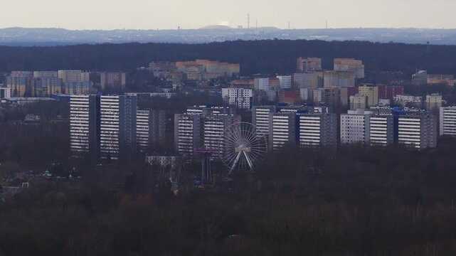 Aerial telephoto pan shows Katowice with leafless parkland, mid rise flats, the Legendia Ferris wheel, and distant chimneys under an overcast daytime sky.