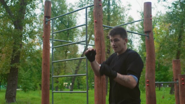 white man boxing outdoors in park performing focused shadowboxing and punch drills near wooden calisthenics bars, wearing black gloves and athletic gear, calm overcast sky, green trees, gritty