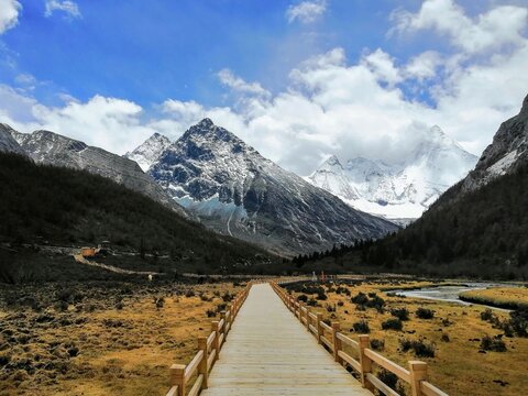 Wooden Boardwalk Leading to Snow-Capped Mountains in Yading Nature Reserve, Sichuan, China. Majestic alpine landscape with clear blue sky and white clouds.