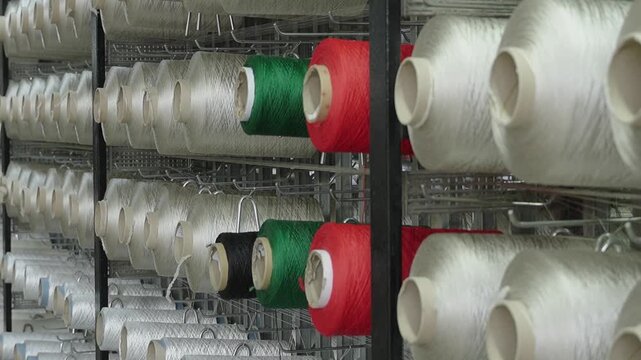 Rows of white and colored industrial yarn bobbins on a metal rack in a textile manufacturing factory.