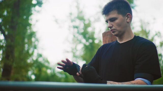 white man tying wraps near fence, wooden posts and green trees surround tattooed arms, serious expression and cinematic shallow depth, last minute ritual before workout or bout, quiet determination