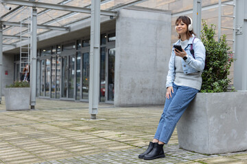 Asian woman 20s sitting on planter in plaza, denim, headphones, bag, holding phone, copy space © wavebreak3