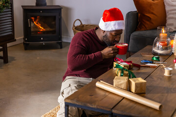 Fototapeta premium African American man sipping red mug in living room wearing Santa hat while wrapping gifts