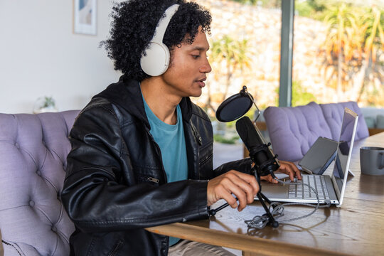 African American man sitting at wooden table using silver laptop and speaking into condenser mic