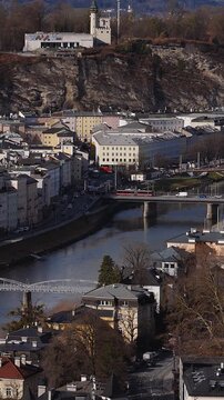 Aerial view of Salzburg, Austria, with Salzach River, baroque Old Town, clock tower, Monchsberg cliffs, Museum der Moderne, tram and cars on bridge, winter light.