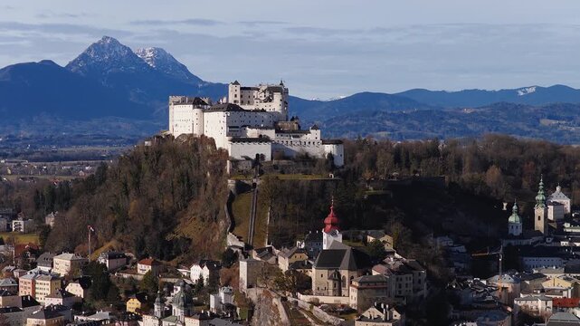 An aerial view over Salzburg, Austria, shows Hohensalzburg Fortress, Salzburg Cathedral domes, a red topped steeple, Untersberg, and Alpine ridges.