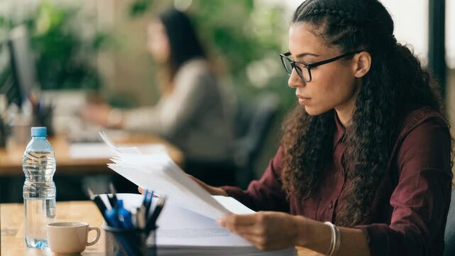 Medical coder organizing printed referrals on a desk highlighting hands sorting documents while the office environment fades softly behind.