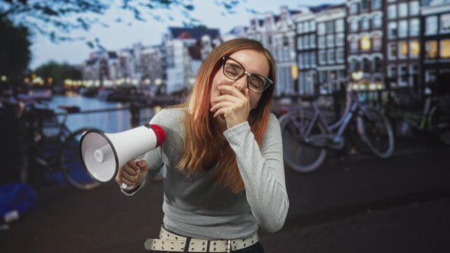 Woman redhead holding megaphone while covering mouth with hand on canal street lined with bicycles and buildings; playful protest.