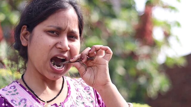 ​A smiling young Indian woman using a traditional Babool (Acacia) herbal twig (datun) for oral care in a lush green outdoor garden.