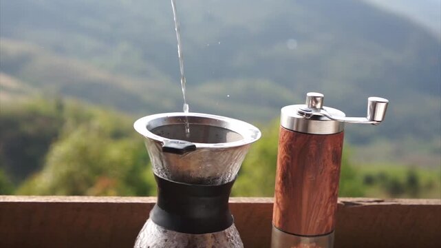 Freshly brewed coffee setup, showcasing a manual grinder and dripper with a scenic outdoor background. A stream of water is pouring, the aromatic smell of the freshly brewed coffee fills the air.