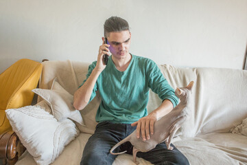 Fototapeta premium A young man sits on a couch holding his phone. A cat sits beside him on the couch
