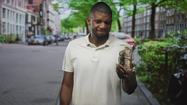 Man holding a glass jar of folded dollars while grimacing on a cobbled city street lined with parked cars and trees; financial frustration.