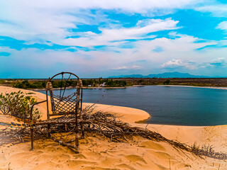 Secluded tropical oasis with sand dunes, a dark water lagoon, and distant mountains under a cloudy sky. © Oscar