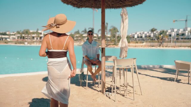 Stylish woman in swimsuit and hat walk on sandy beach towards smiling man sitting under tiki umbrella, enjoy romantic summer getaway at luxurious waterfront resort.Vacation concept for tourism