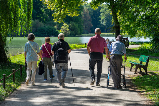 Zwei &auml;ltere M&auml;nner, und andere Senioren gehen entspannt im Park spazieren -  Sohn und Opa mit Rollator