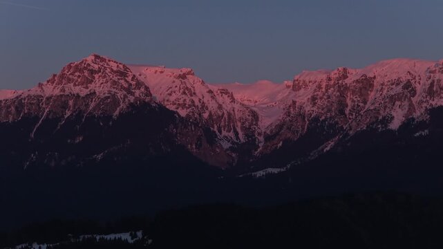 Stunning intense red alpenglow on the snowy Bucegi Mountains peaks of Scara and Omu at sunset, 70mm medium tele aerial pan, cinematic 4K