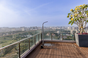 Fototapeta premium Rooftop terrace featuring a wooden deck, glass railing and potted flowering tree with sweeping city skyline and high-rise buildings in the distance. Terrace garden with city view