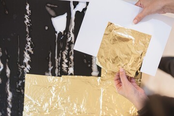 Hands gently apply shimmering gold leaf to a stunning piece of luxury furniture in a vibrant artist’s studio, surrounded by creative chaos and rich colors © Damian