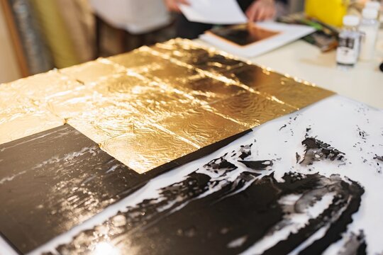 In a serene studio, an artist applies gold leaf to a striking artwork, merging luxurious gold with bold black and white, highlighting a detailed creative process