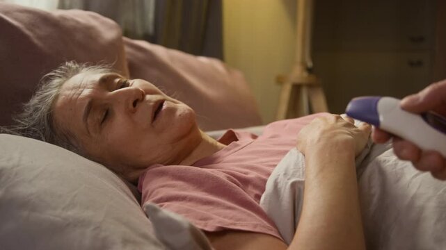 Grey-haired senior lady lying in bed sick with high fever, unknown man checking her temperature with infrared thermometer and putting cold compress on her forehead