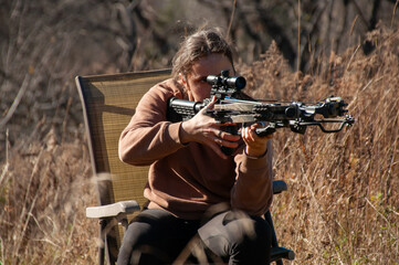 Fototapeta premium A young woman sitting in a chair, carefully aims her crossbow getting ready to shoot. She practices outside in a grass field during a sunny day in autumn.