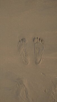 Vertical video of an ocean wave washing away bare footprints in wet beach sand
